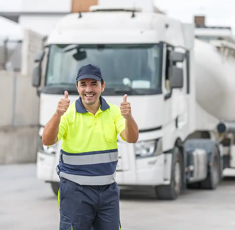 A man in a neon yellow safety shirt and blue cap stands in front of a large white lorry, smiling and giving two thumbs up. The setting appears to be an industrial or car park area.