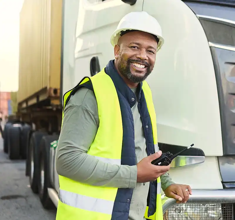 A man wearing a white hard hat and yellow safety vest stands in front of a lorry, smiling and holding a walkie-talkie. A shipping container is visible on the lorry behind him.