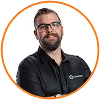 A man with short dark hair, a beard, and glasses is wearing a black collared shirt with a small logo related to transport compliance. He is smiling slightly and has his arms crossed. The image has an orange circular border.